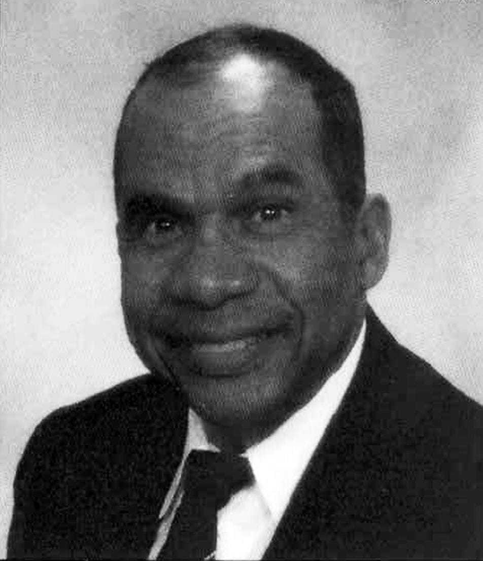 A black and white headshot of Omego Ware in a suit and tie.